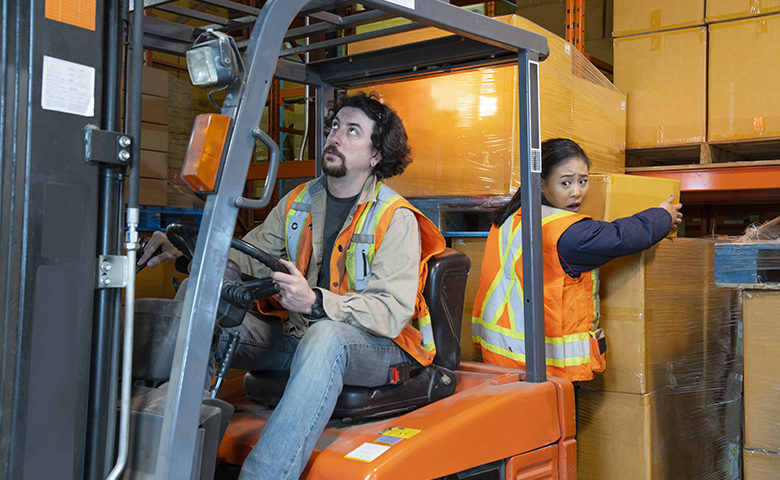 An industrial warehouse workplace safety topic. A worker in the danger zone, working behind a forklift carrying a load.